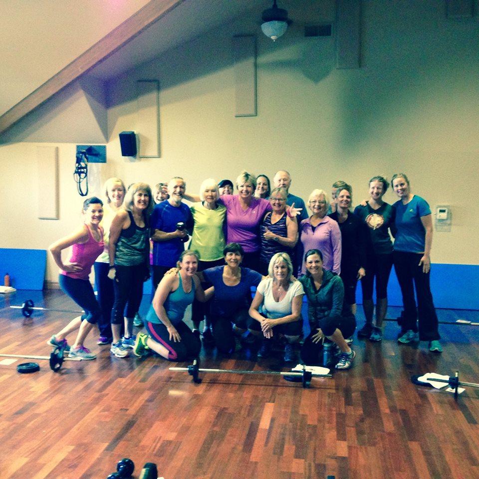 A smiling, multi-generation group of club members pose together in a bright fitness studio after a strength class, with barbells and weights on the wooden floor highlighting their shared commitment to health, wellness, and community.